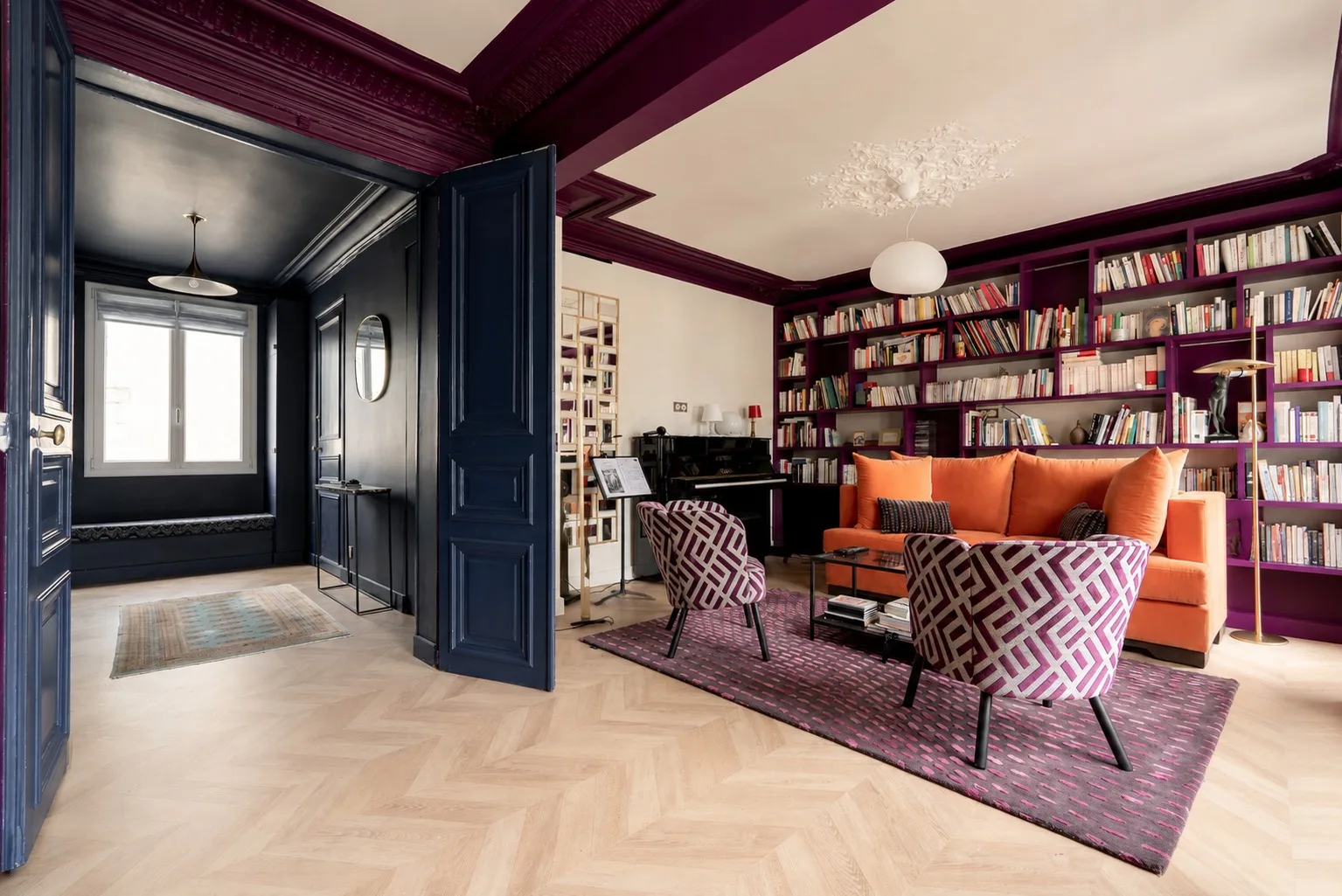 Una sala de estar con estanterías, asientos naranjas y sillones estampados alrededor de una alfombra.