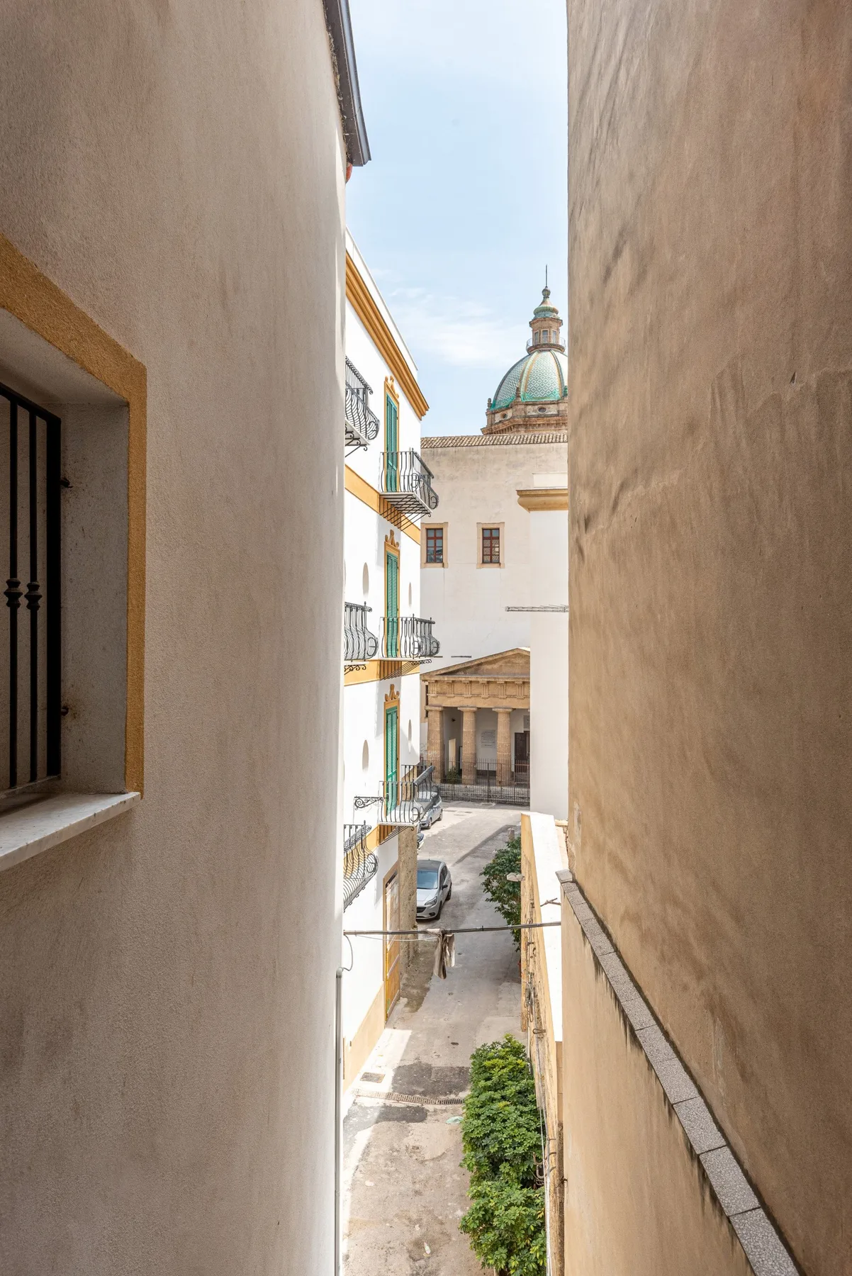 A narrow outdoor passage between buildings overlooks a sunlit courtyard and domed tower.