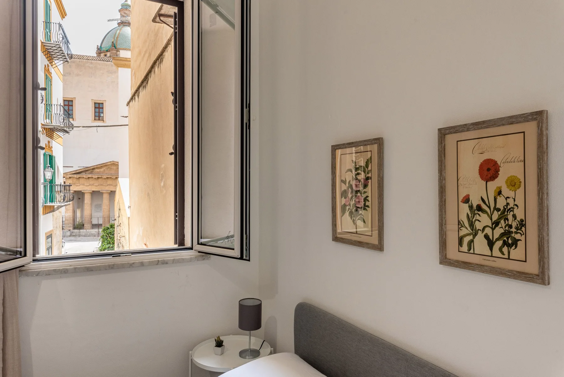 A bedroom with a bed, framed botanical prints, and an open window.