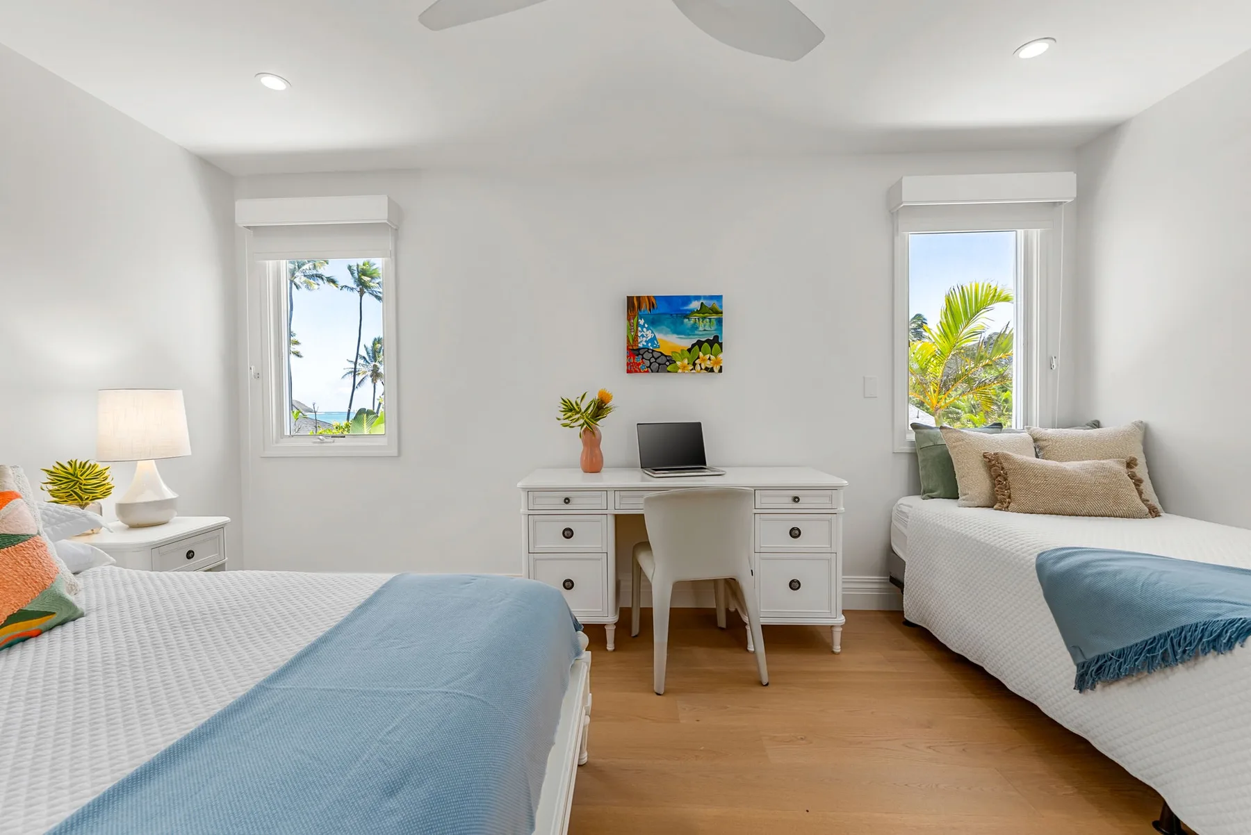 Un dormitorio luminoso con dos camas y un escritorio bajo un cuadro colorido.