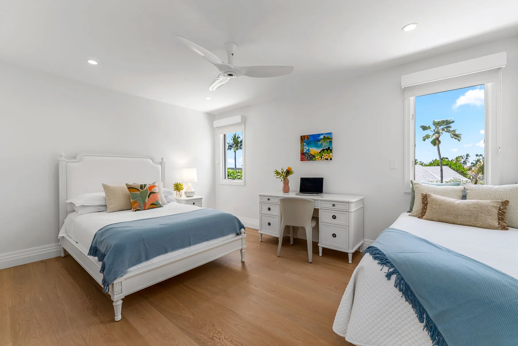 Un dormitorio luminoso con dos camas y un escritorio entre las ventanas.