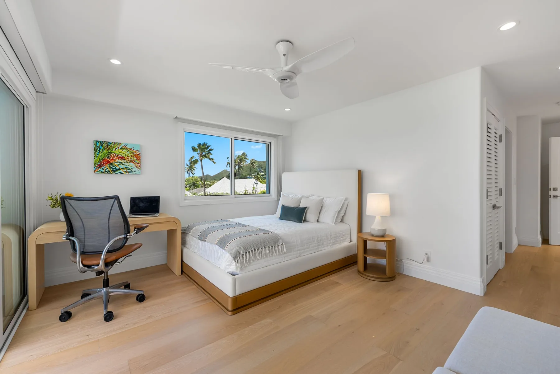 Un dormitorio luminoso con cama, escritorio y ventana con vista a palmeras.