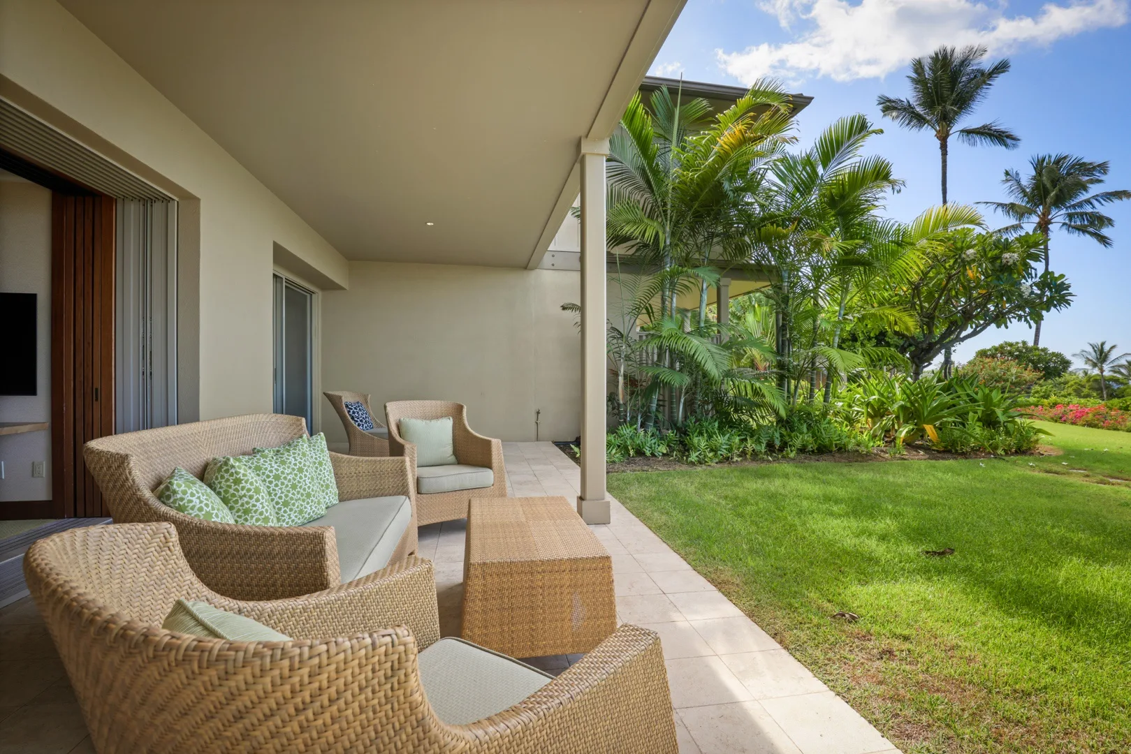 Un patio cubierto con asientos tejidos da a un césped verde y plantas tropicales.