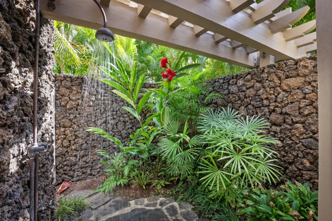 Una ducha exterior rodeada de plantas tropicales y muros de piedra.