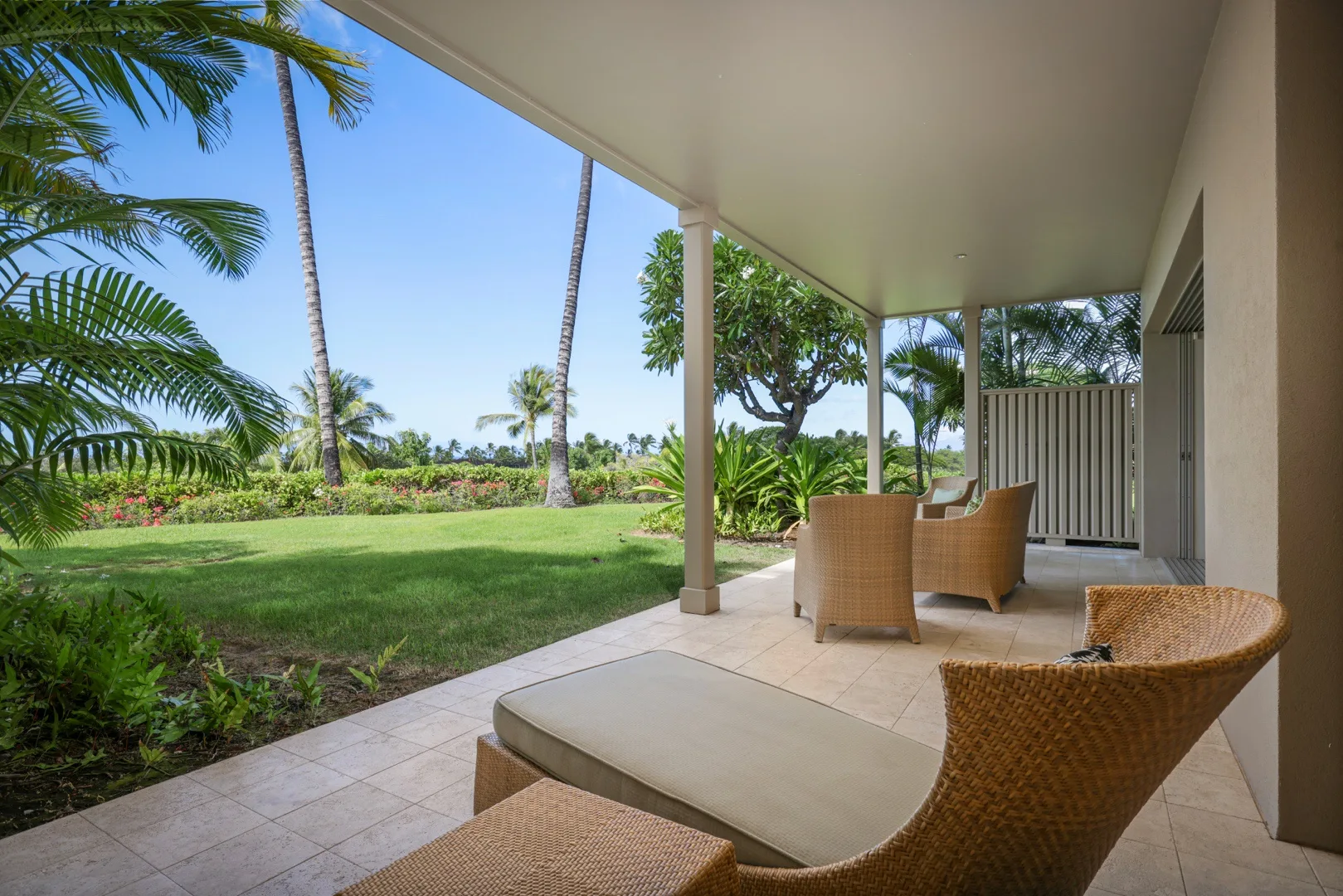 Un patio cubierto con asientos tejidos da a un césped verde y plantas tropicales.