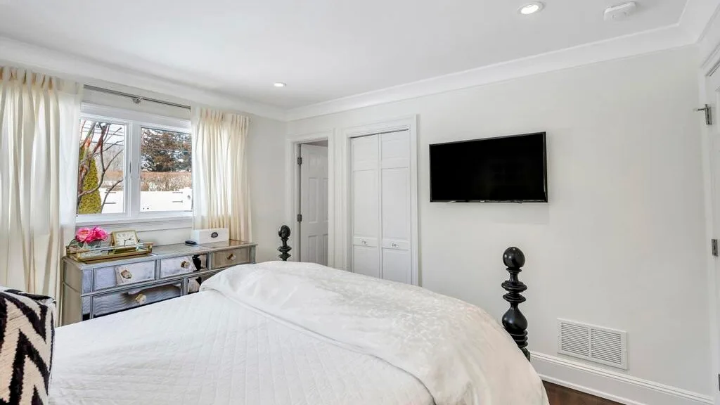 A bright bedroom with a white bed, dresser, and wall-mounted television.