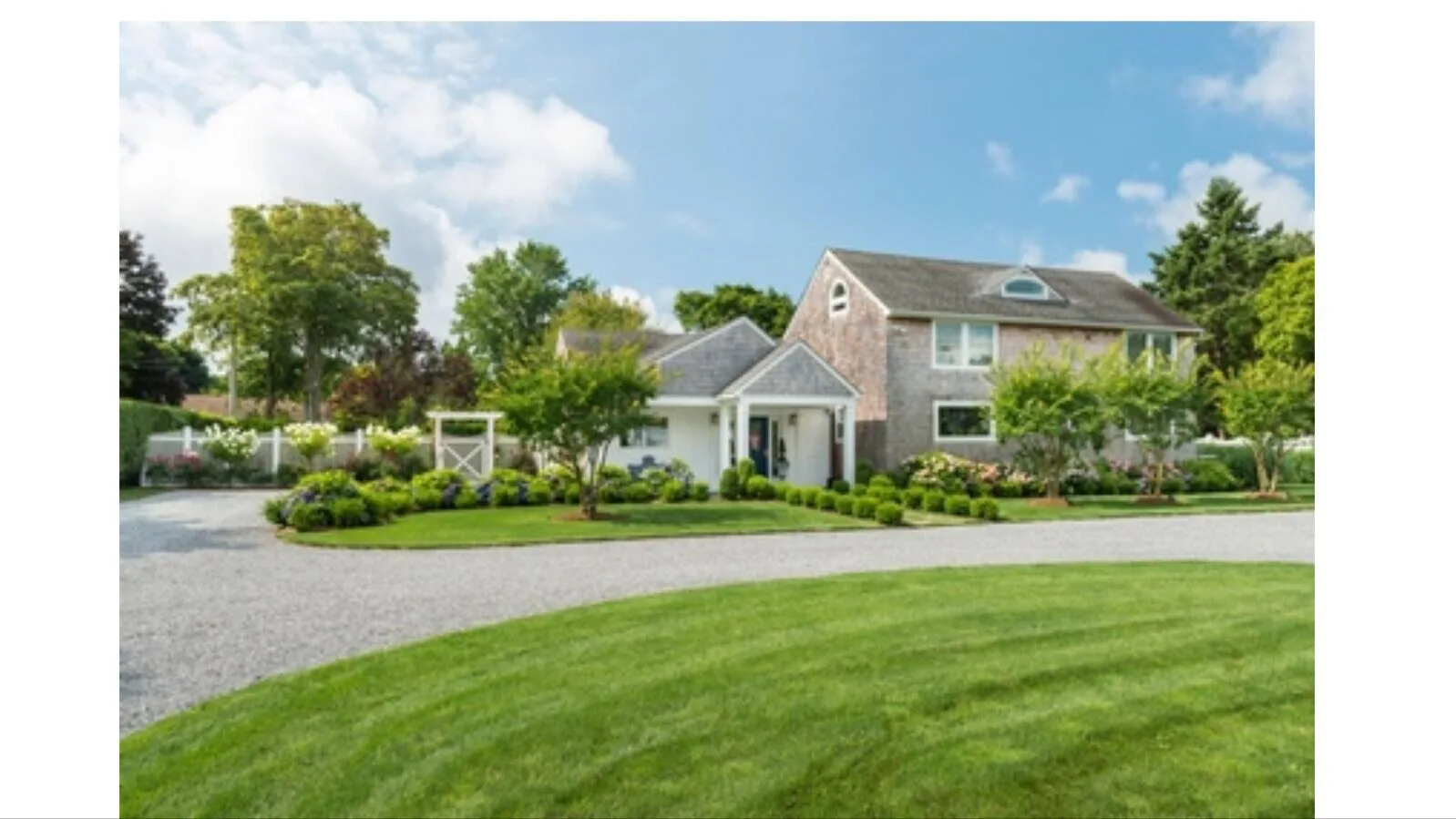 A large house stands behind a landscaped garden and curved gravel driveway.