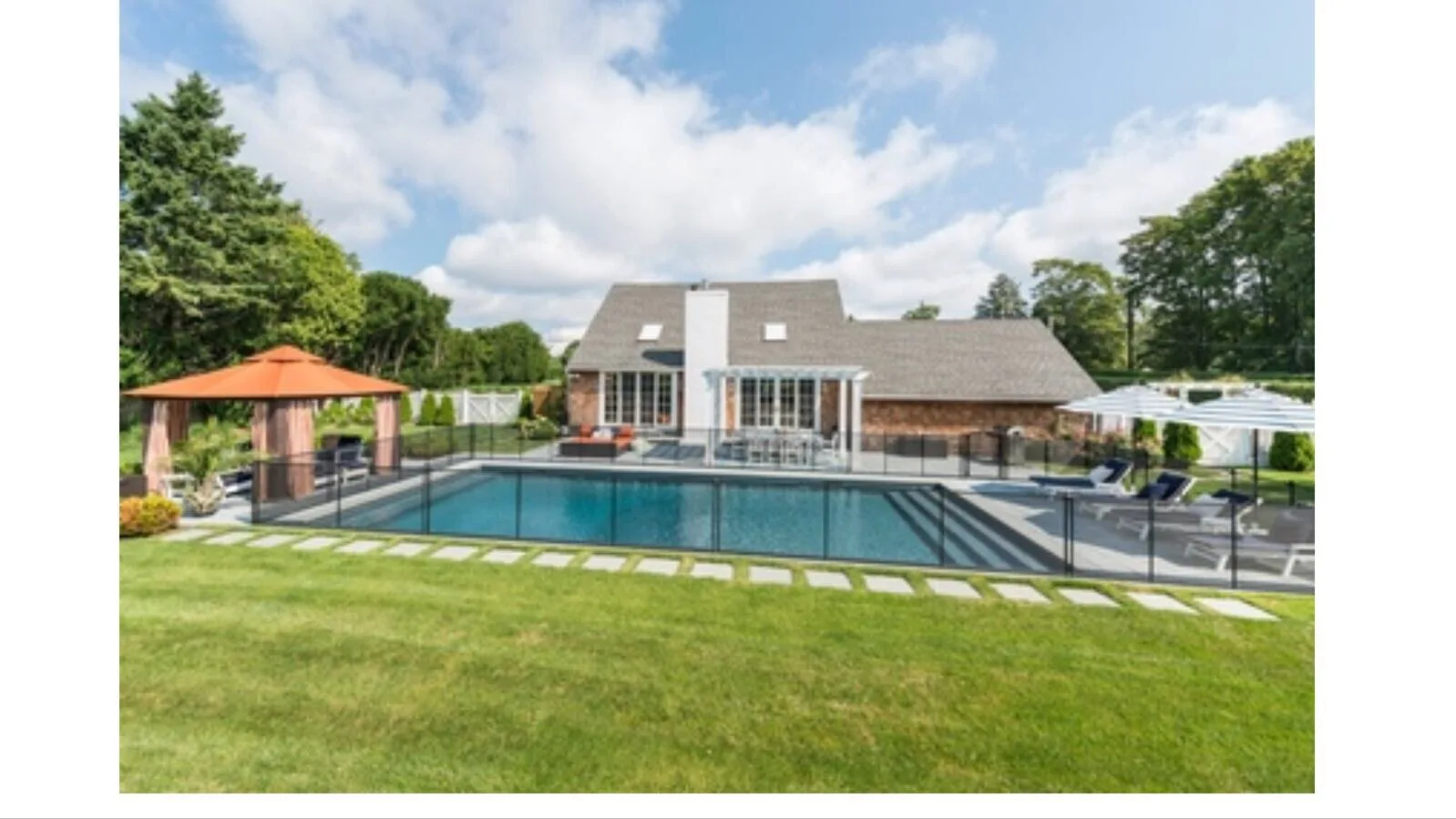 A backyard swimming pool sits beside a house, with a gazebo and lawn.