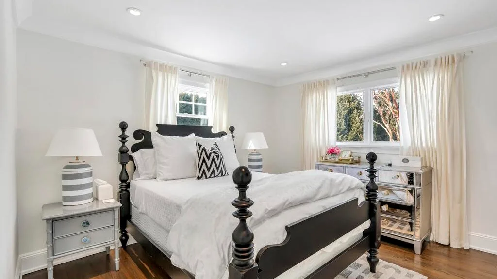 A bright bedroom with a dark bed, white bedding, and windows.