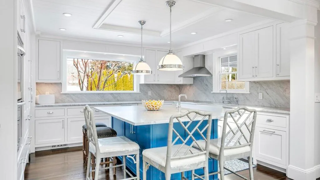 A bright white kitchen features a blue island with seating and pendant lights.