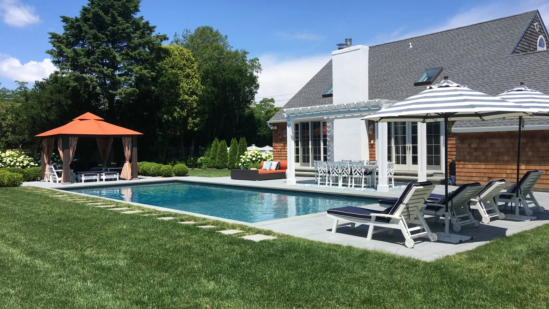 Una piscina junto a una casa, con asientos de descanso y sombrillas.