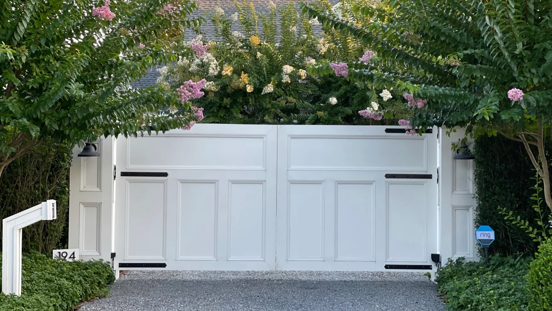 A white driveway gate is framed by leafy greenery and flowering branches.