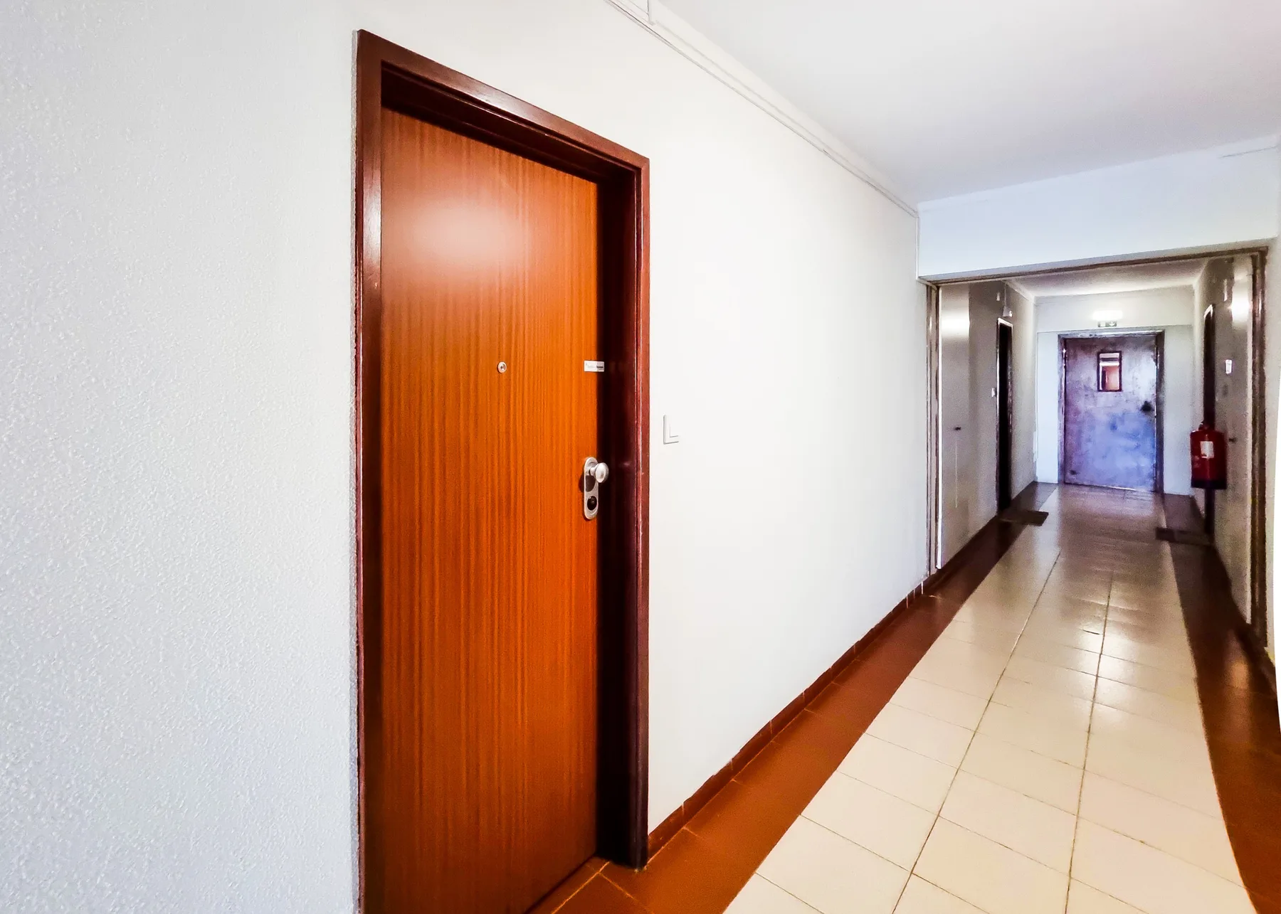 A tiled hallway with a wooden apartment door and a fire extinguisher.