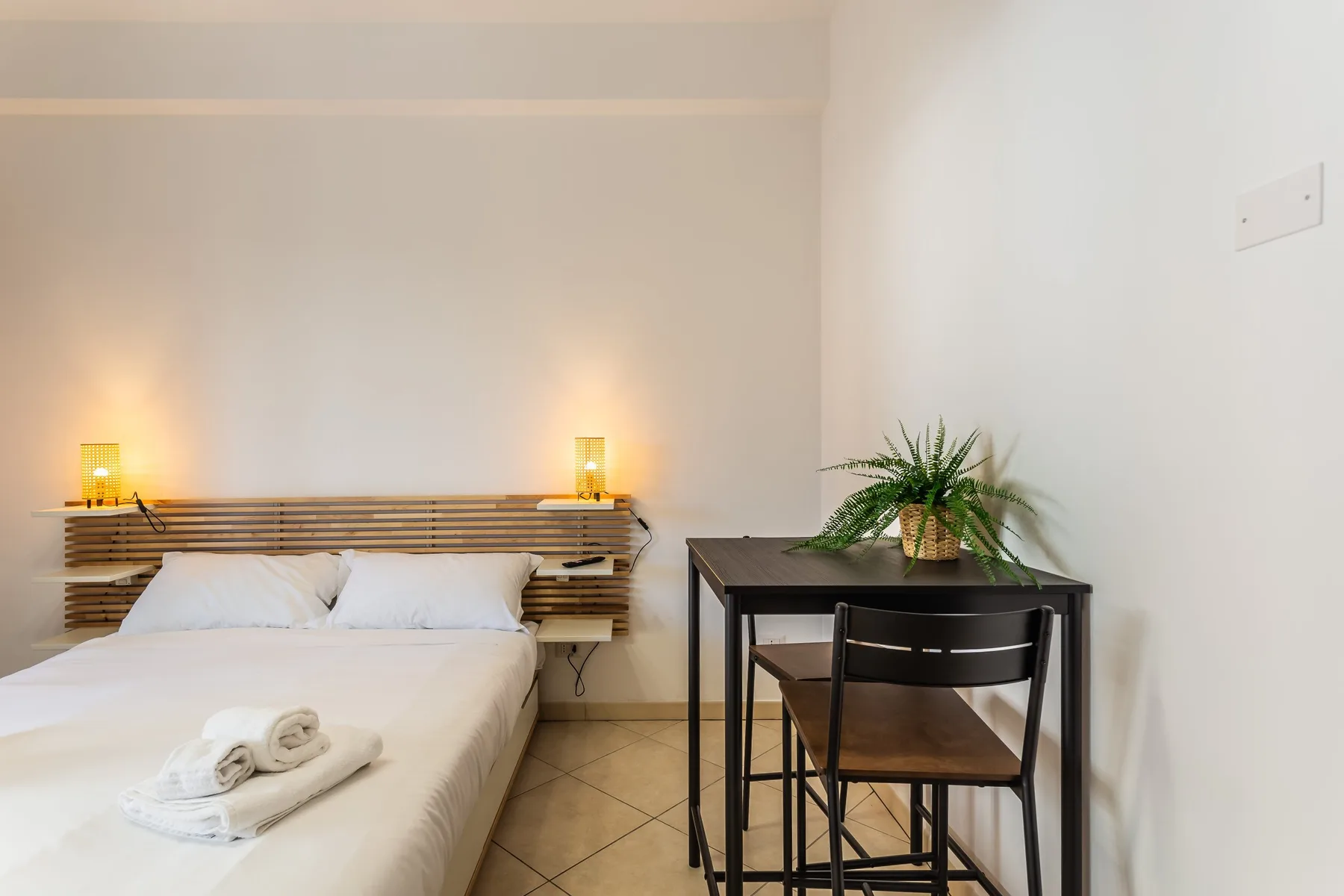 A bedroom with a bed, bedside lights, and a small desk with a plant.
