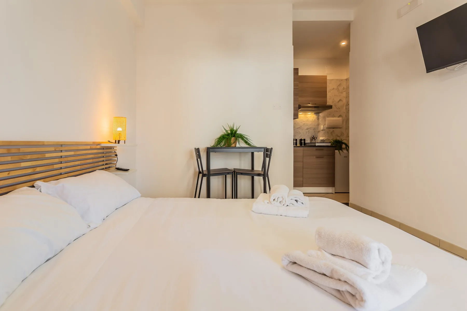A bedroom with a bed, a small table, and an open kitchenette in the background.