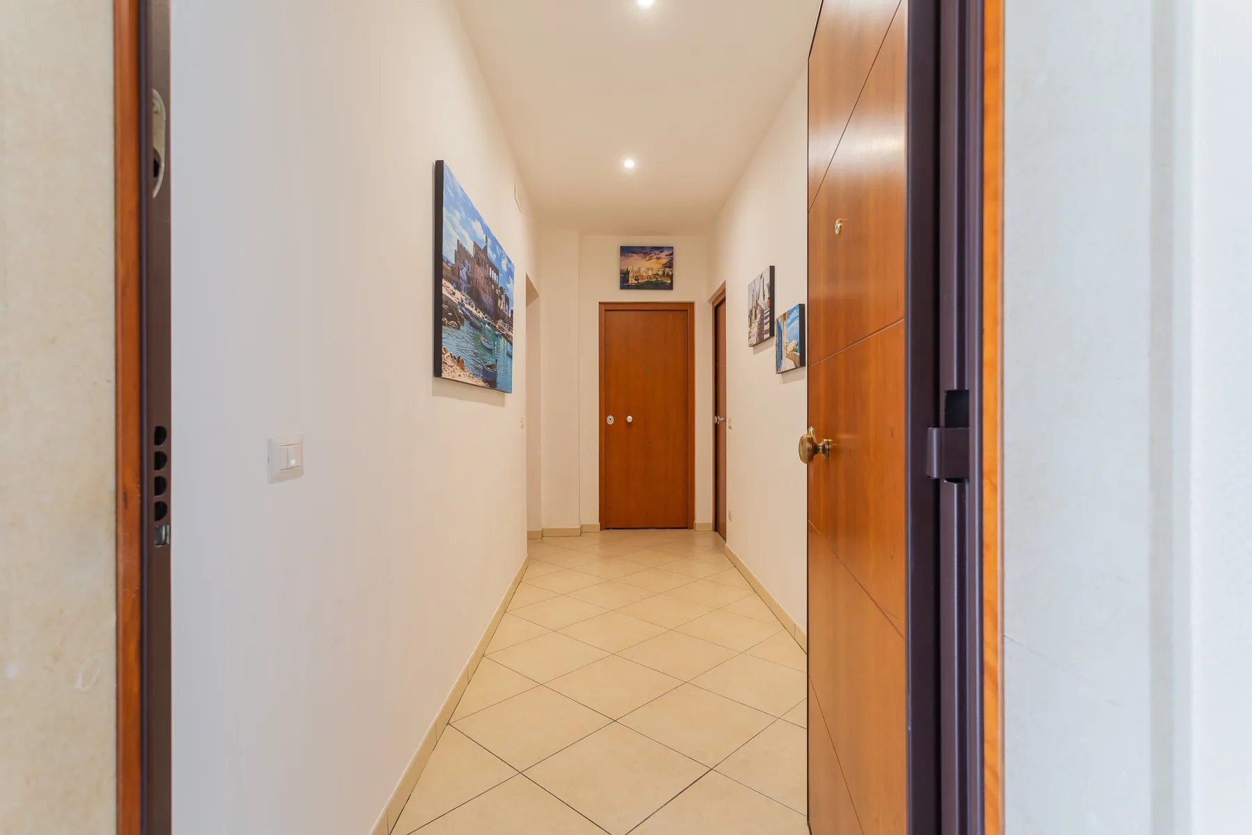 A bright hallway with tiled flooring, wall art, and a wooden door.