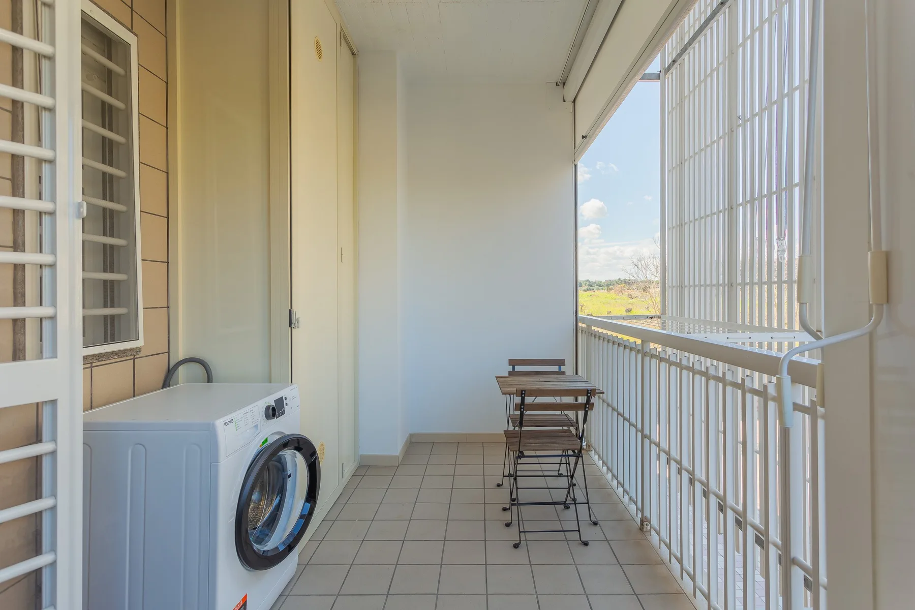 A narrow balcony has a washing machine and a small table with chairs.