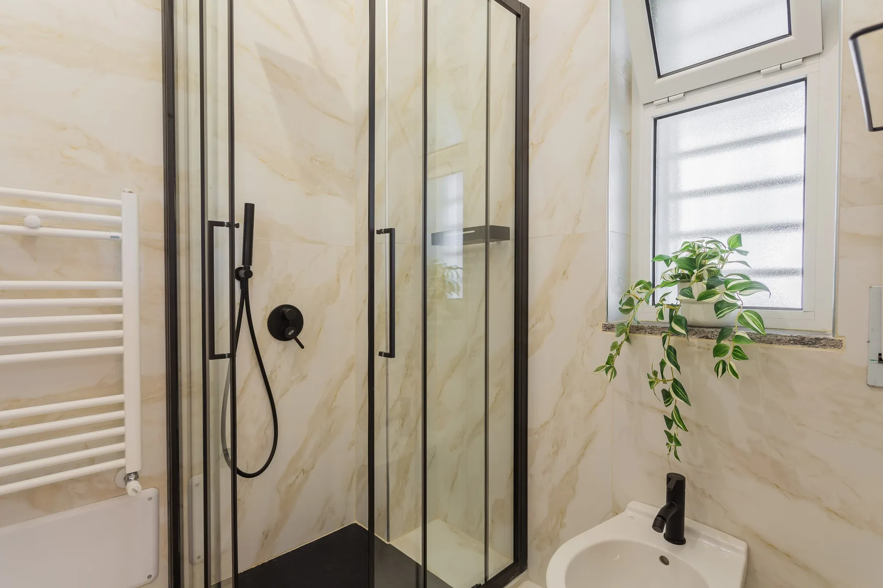 A bathroom with a glass shower, sink, and leafy plant by the window.