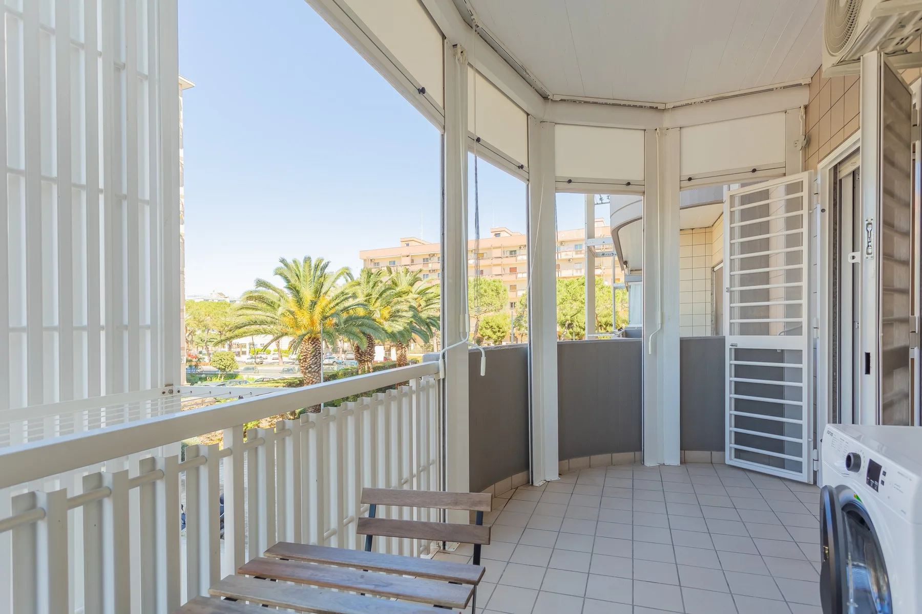 An enclosed balcony with tiled flooring and views of palm trees and nearby buildings.