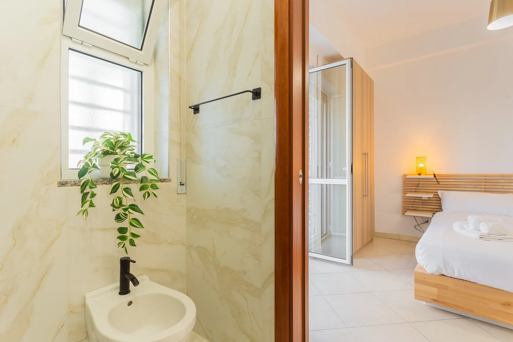 A bathroom sink and mirror open onto a bedroom with a bed and desk.
