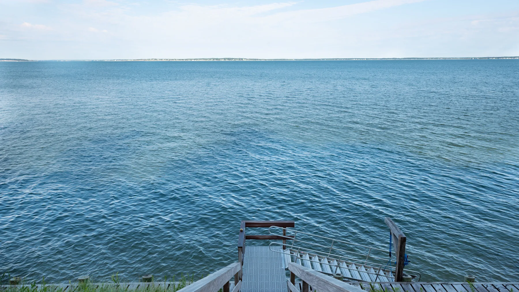 A wooden pier extends over calm blue water beneath a partly cloudy sky.