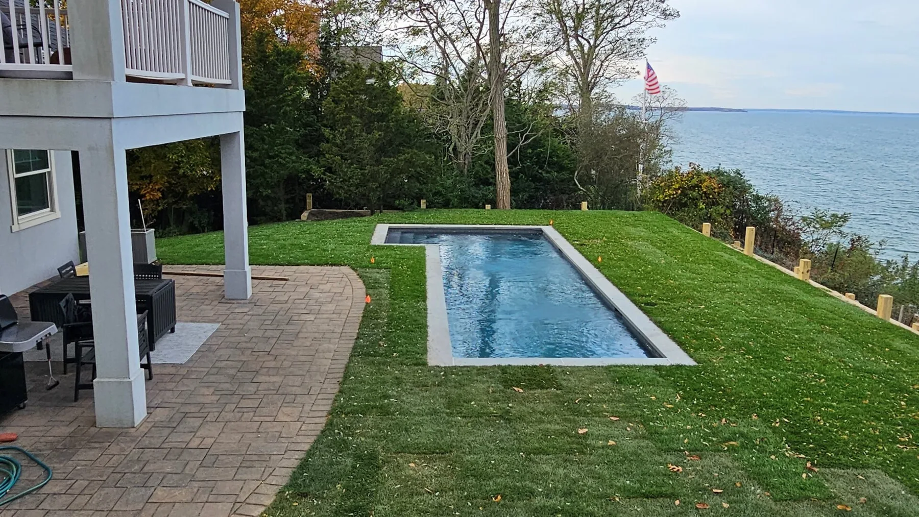 A backyard pool beside a paved patio, with a lawn and water in the background.