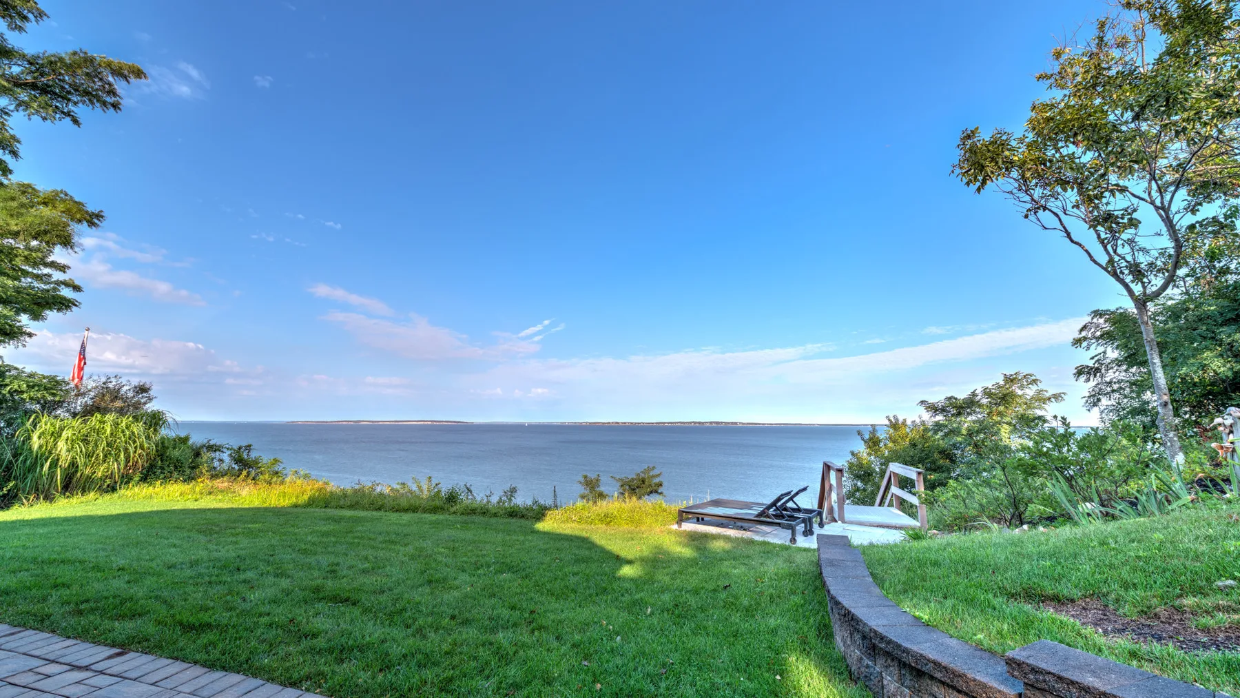 A grassy waterfront lawn overlooks calm water, with a small seating area nearby.