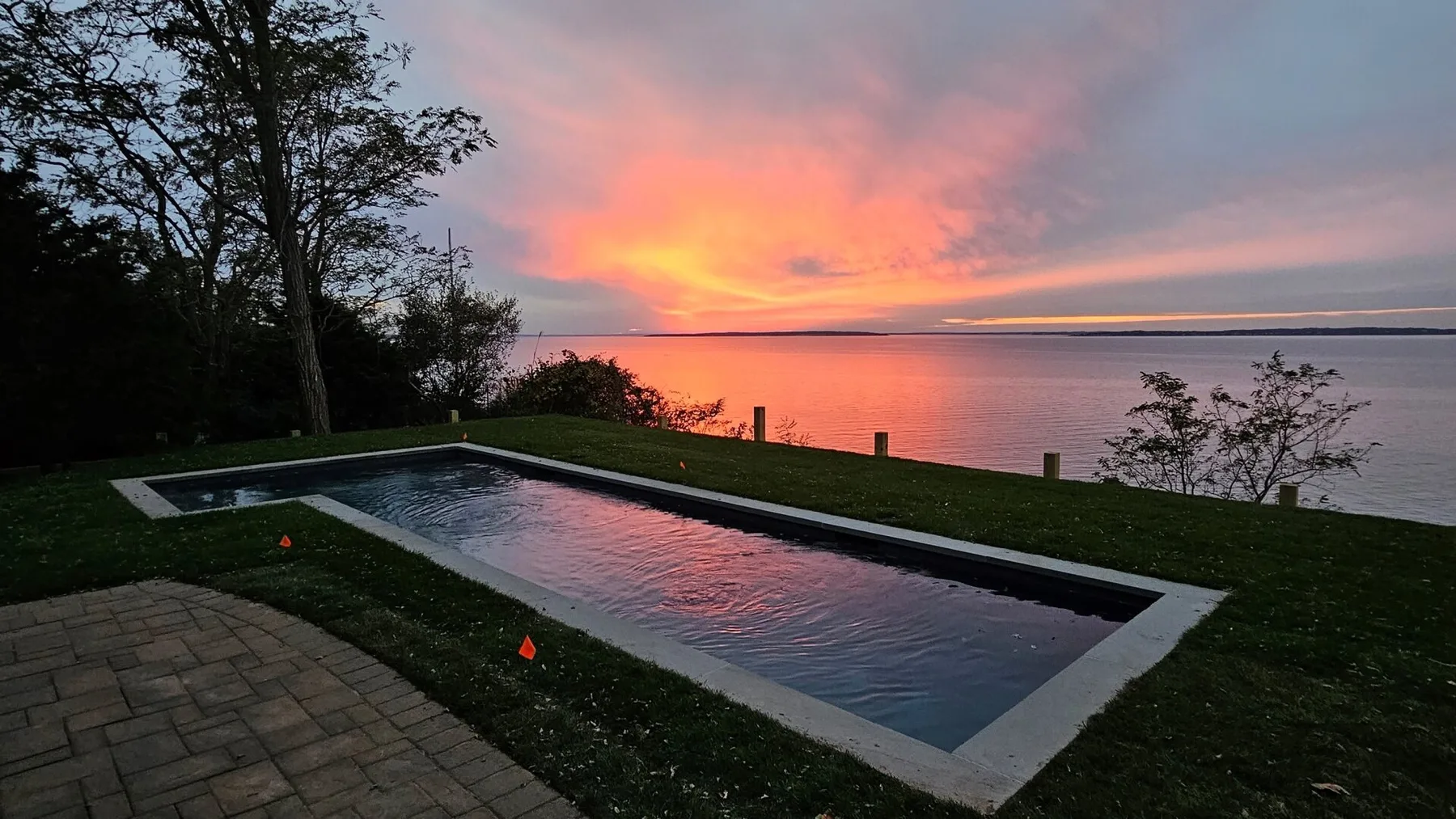 A rectangular pool overlooks calm water beneath a vivid sunset sky.