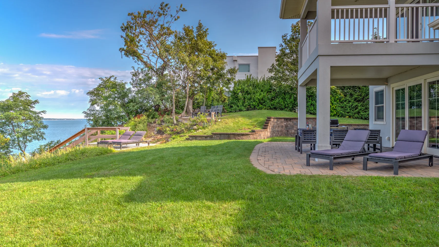 A waterfront lawn beside a patio with outdoor seating and trees.