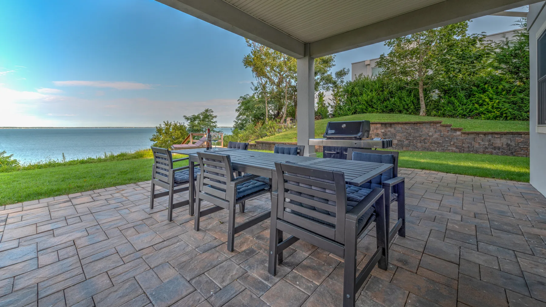 A covered patio overlooks a waterfront, with an outdoor dining table and seating.