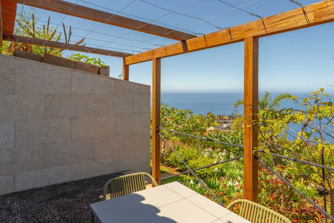 An outdoor dining area beneath a wooden pergola overlooks the sea and greenery.