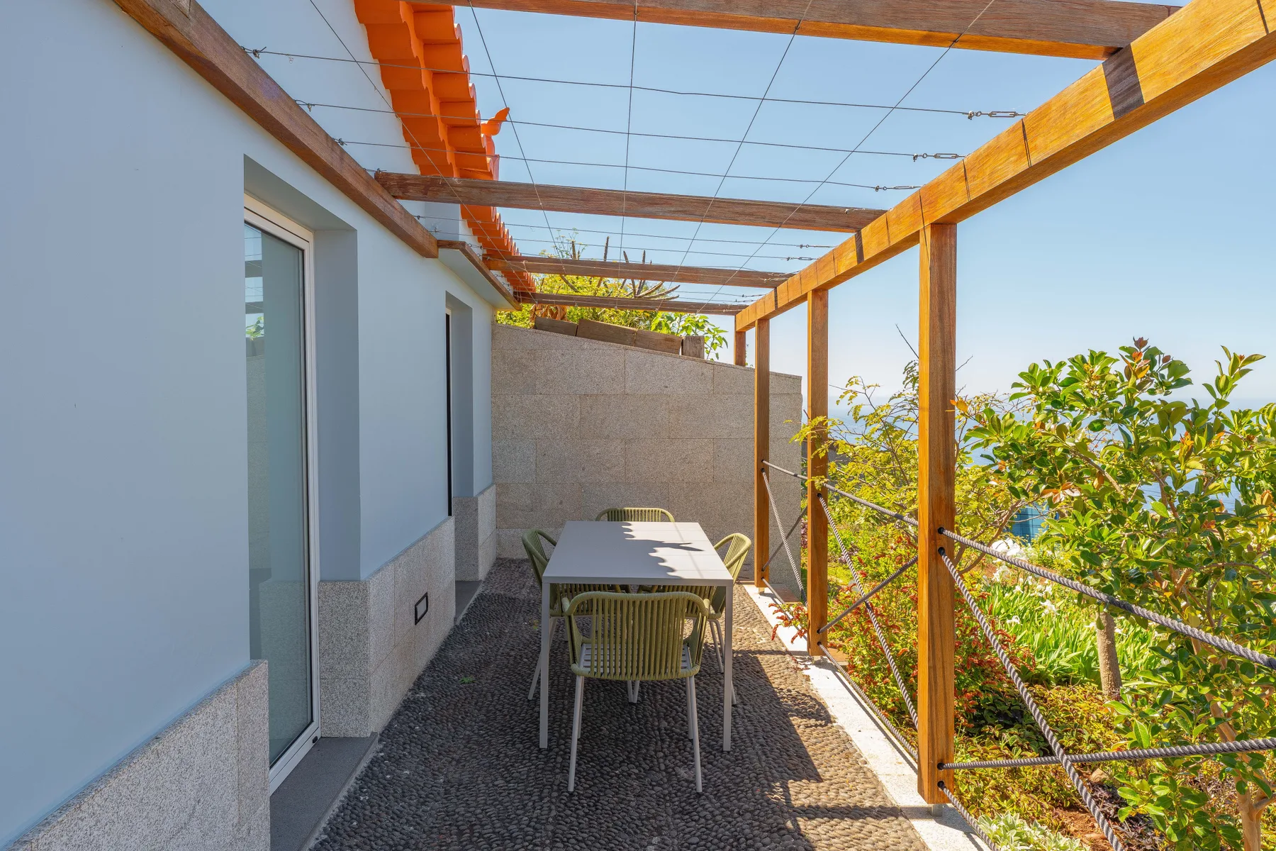 An outdoor dining area sits beneath a wooden pergola beside a garden.