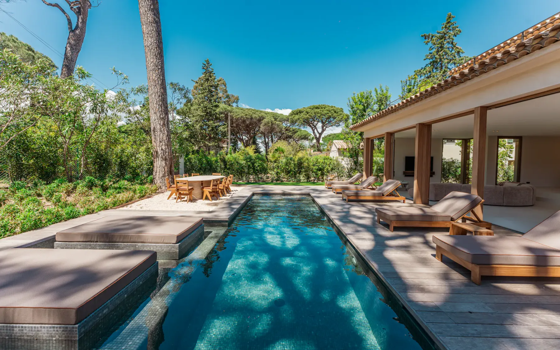 Vista desde un extremo de la piscina al aire libre, con una villa a la derecha, cómodas tumbonas y un jardín bien cuidado con árboles a la izquierda.