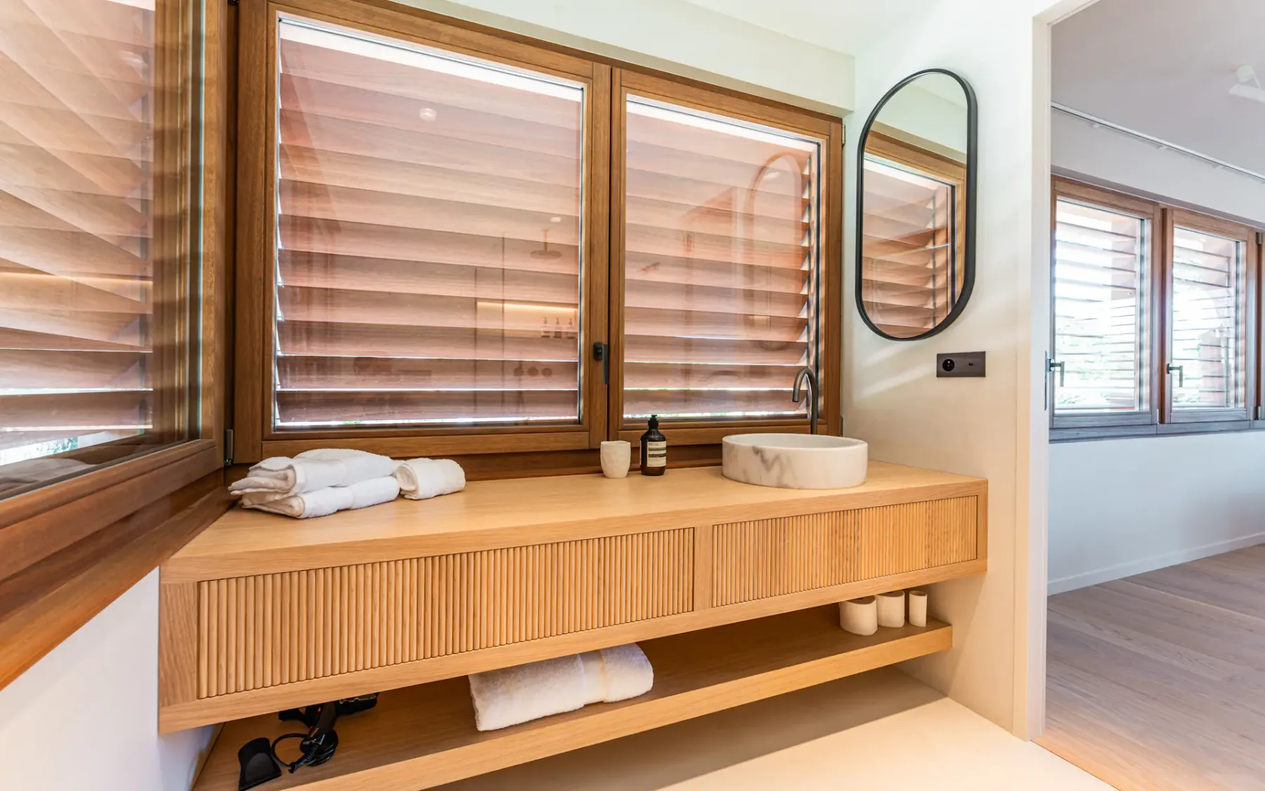 Mueble de baño grande de madera con un lavabo redondo a la derecha, un espejo montado en la pared y ventanas con persianas de madera cerradas.