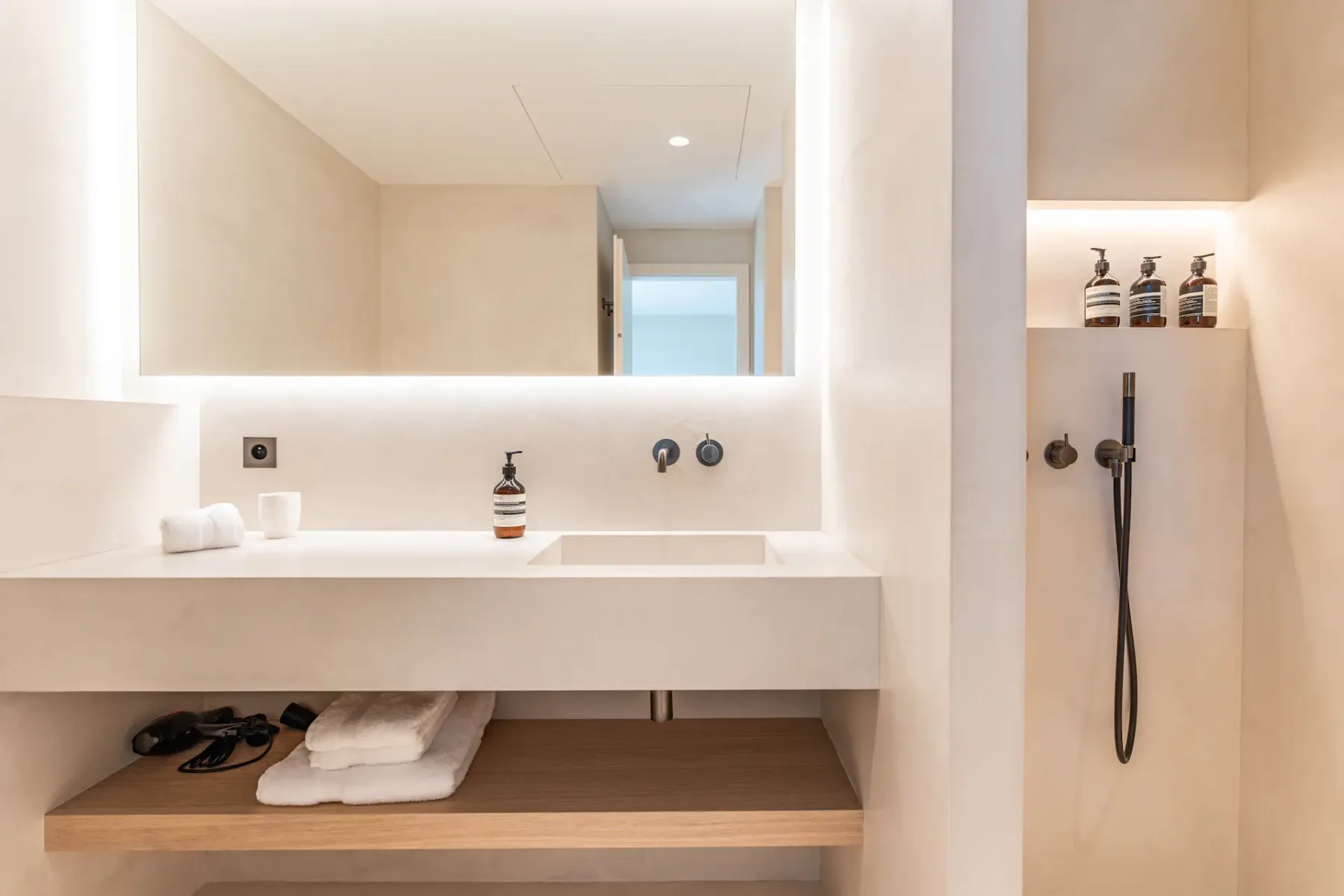 Baño moderno y limpio con un lavabo blanco, un estante de madera debajo, un espejo grande encima y una ducha blanca a la derecha.