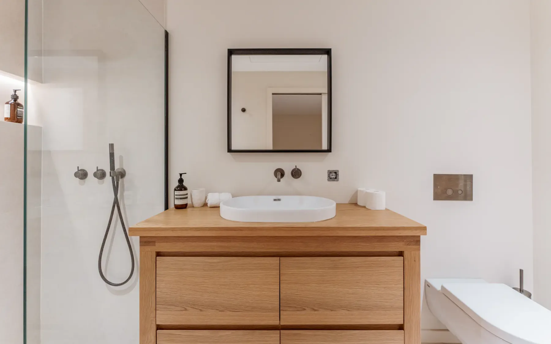 Vista de un mueble de baño de madera con un lavabo y un espejo cuadrado montado encima, con un inodoro a la derecha y una ducha con puerta de cristal a la izquierda.