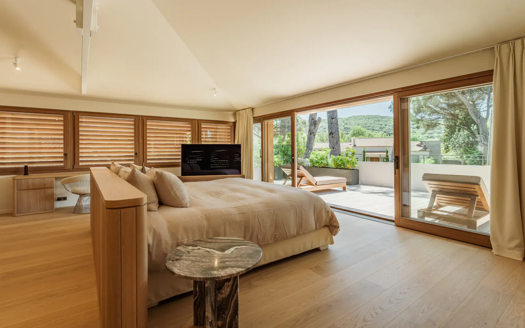 Dormitorio grande con luz natural, una cama doble grande en el centro con cabecero de madera, un televisor de pantalla plana, grandes puertas de cristal abiertas que dan a un exuberante jardín y ventanas con persianas de madera en un lateral.