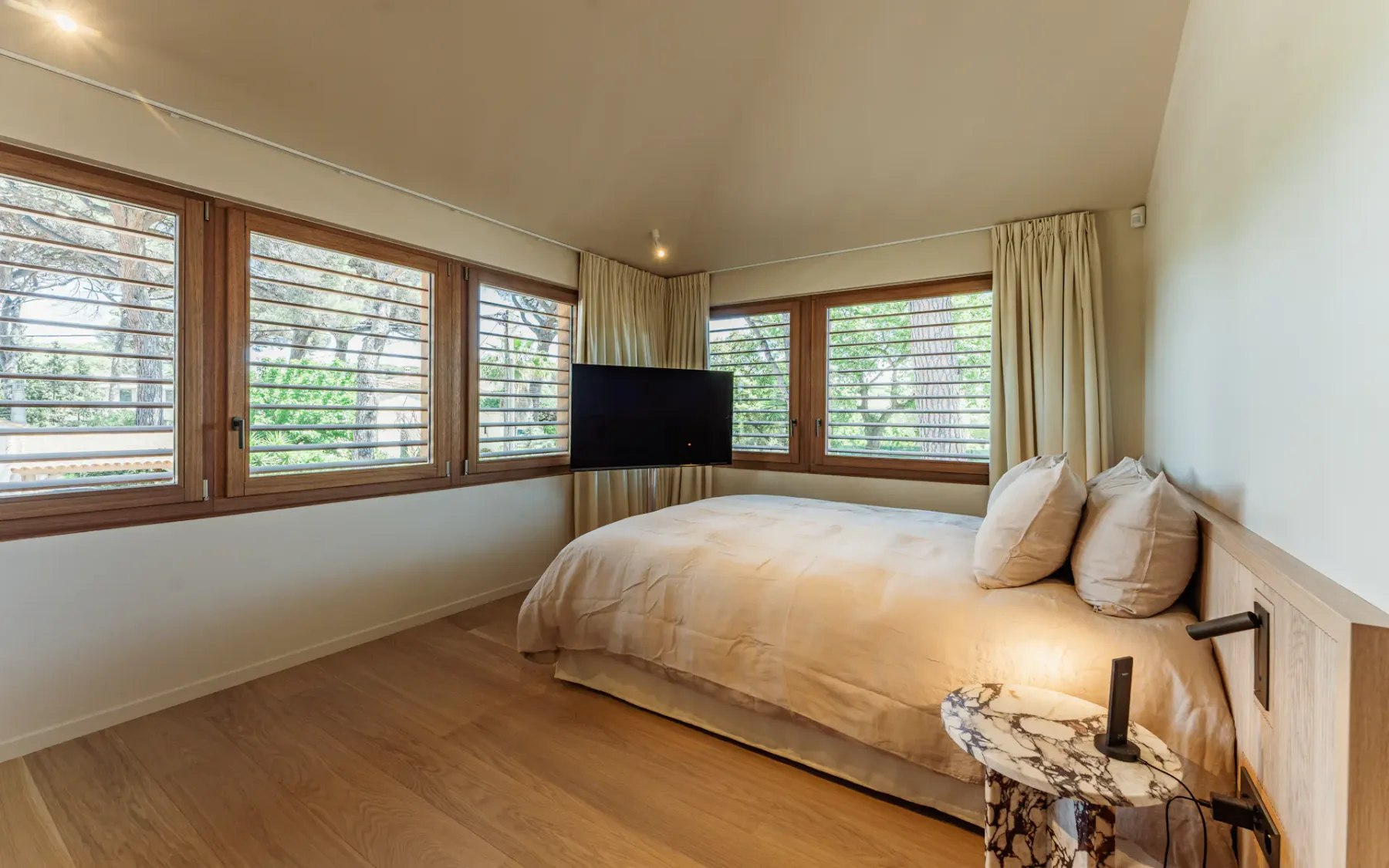 Dormitorio grande y moderno con una cama doble, una mesa de noche con lámpara, suelo y puerta de madera, un televisor de pantalla plana y grandes ventanas con las persianas abiertas que dejan entrar luz natural.