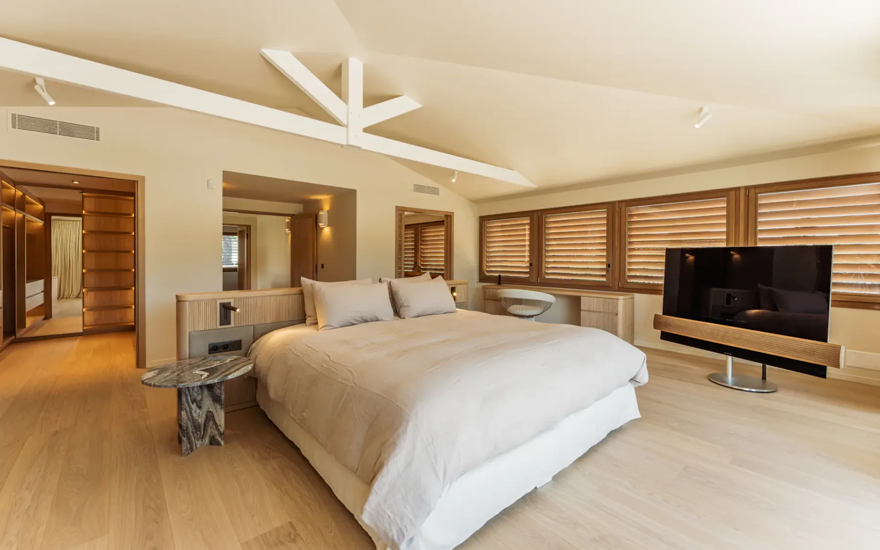 Dormitorio grande con luz natural, una cama doble grande en el centro, ventanas con persianas de madera en un lateral, un televisor de pantalla plana y una puerta al fondo que conduce a un amplio vestidor.
