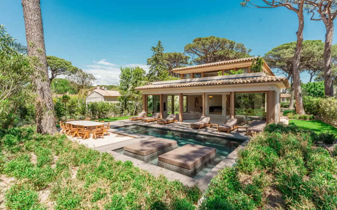 Un patio de villa con una piscina rectangular, asientos al aire libre y vegetación alrededor.