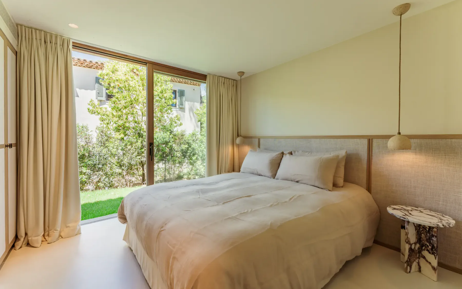 Dormitorio moderno con una cama doble, una lámpara colgante junto a la cama y grandes puertas dobles de cristal que dan a un exuberante jardín.