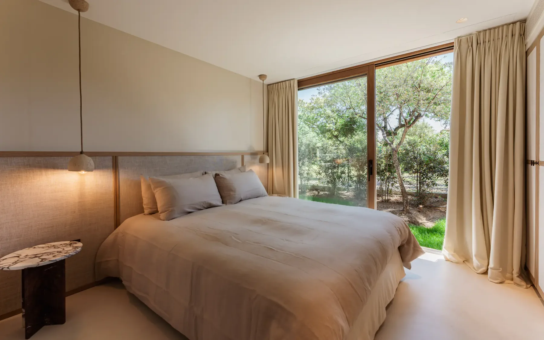 Dormitorio moderno con una cama doble, una lámpara colgante junto a la cama y grandes puertas dobles de cristal que dan a un exuberante jardín.