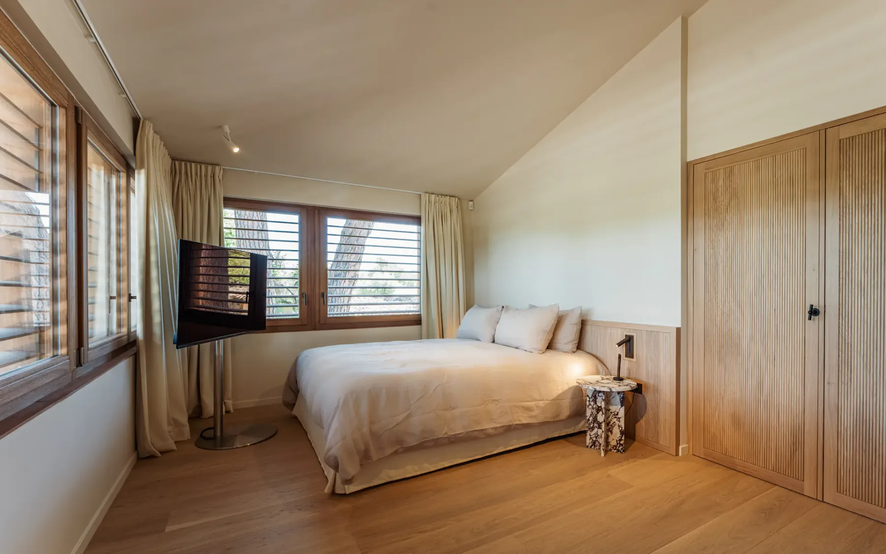 Dormitorio grande y moderno con una cama doble, una mesa de noche con lámpara, suelo y puerta de madera, un televisor de pantalla plana y grandes ventanas con las persianas abiertas que dejan entrar luz natural.