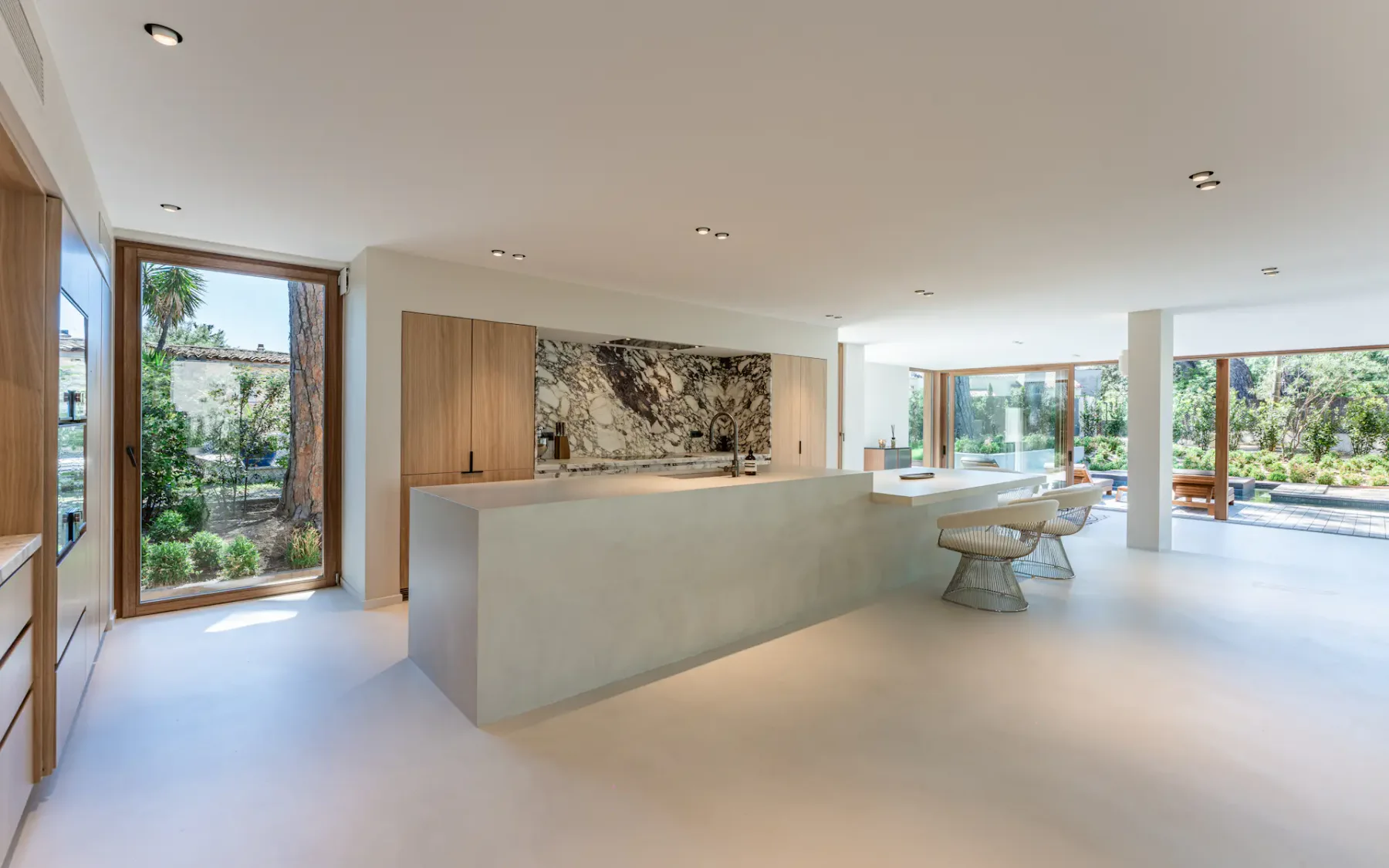 Cocina moderna abierta en una casa contemporánea, con una isla central con barra, taburetes y un revestimiento antisalpicaduras blanco y negro detrás, entre ventanas luminosas que dejan ver abundante vegetación en el exterior.