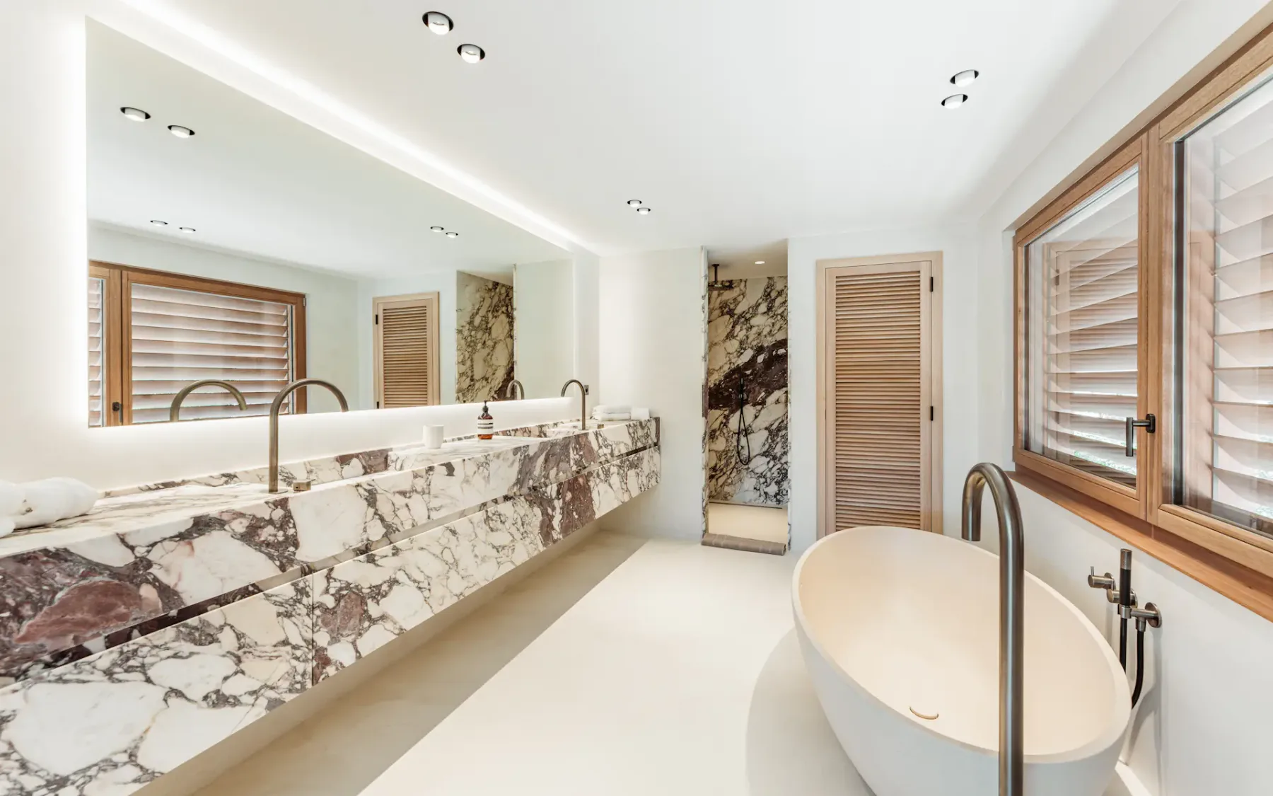 Baño moderno y luminoso con un lavabo de mármol con grifos altos, una bañera grande y una ducha con detalles de mármol al fondo.