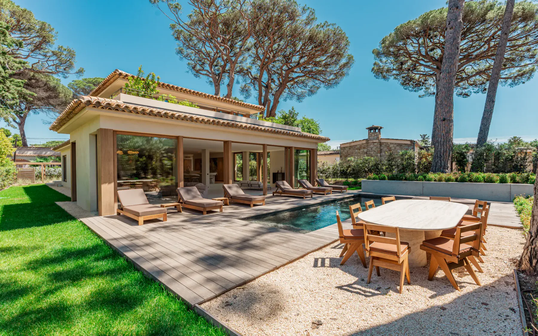 Vista del patio trasero de una villa con césped verde bien cuidado, tumbonas, una piscina y un conjunto de mesa grande y sillas de comedor al aire libre, con árboles altos al fondo.