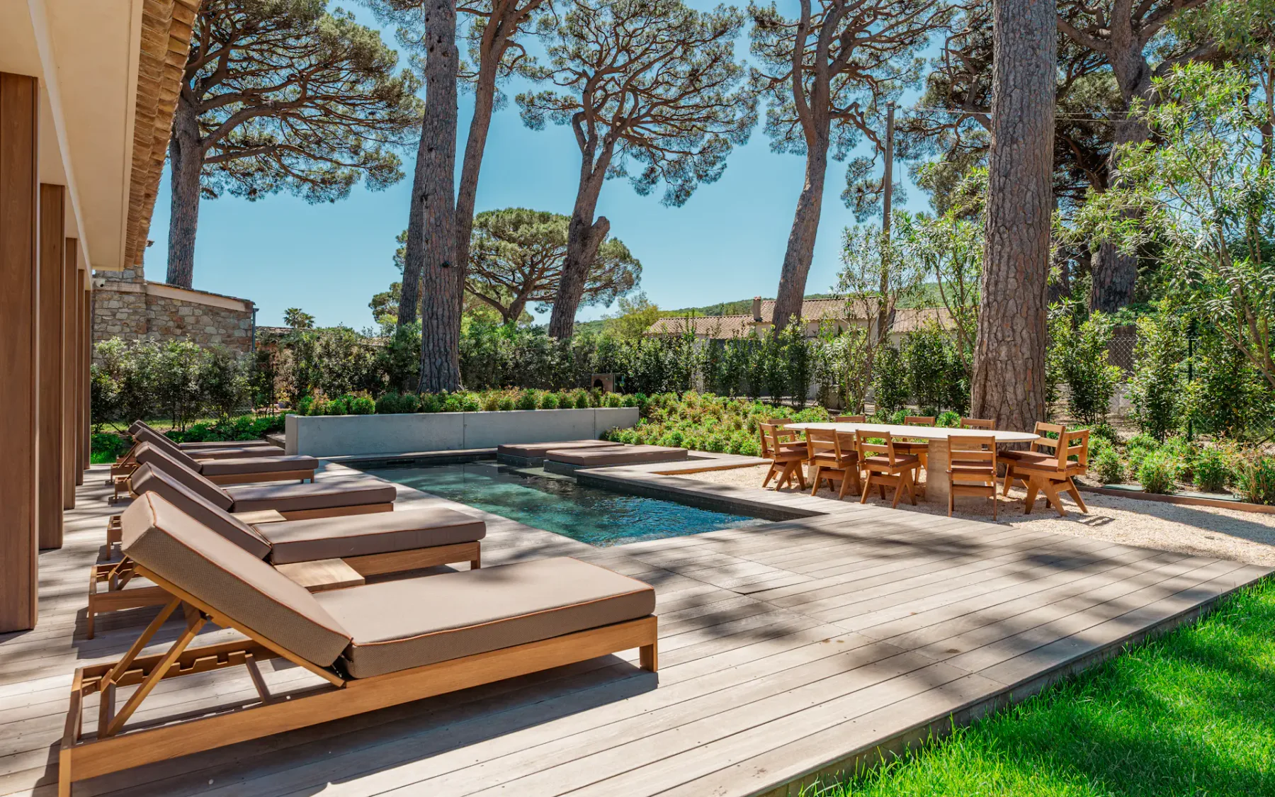 Vista de un jardín impecablemente cuidado con una piscina en el centro, una fila de cómodas tumbonas en la terraza, un conjunto de mesa grande y sillas de comedor al aire libre y un exuberante jardín con árboles altos al fondo.