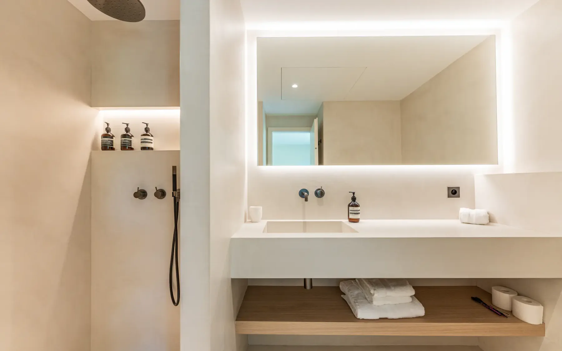 Baño blanco moderno con un lavabo blanco, un estante de madera debajo, un espejo grande retroiluminado y una ducha blanca a la izquierda.