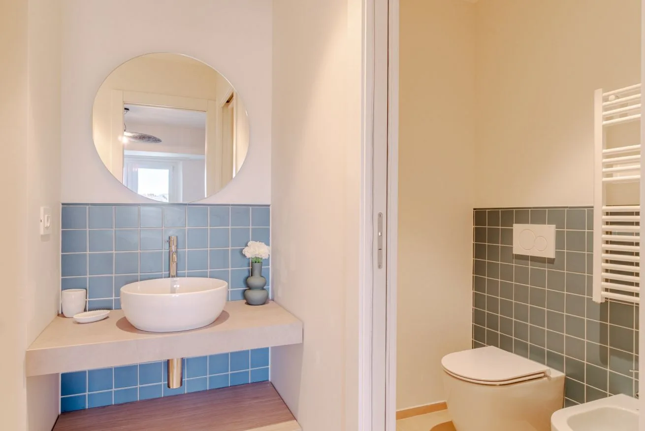 Un lavabo de baño con espejo redondo junto a una zona de inodoro con azulejos.