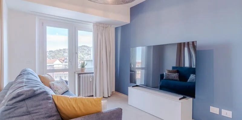 Una sala de estar con asientos, un televisor y ventanas con vistas a edificios cercanos.